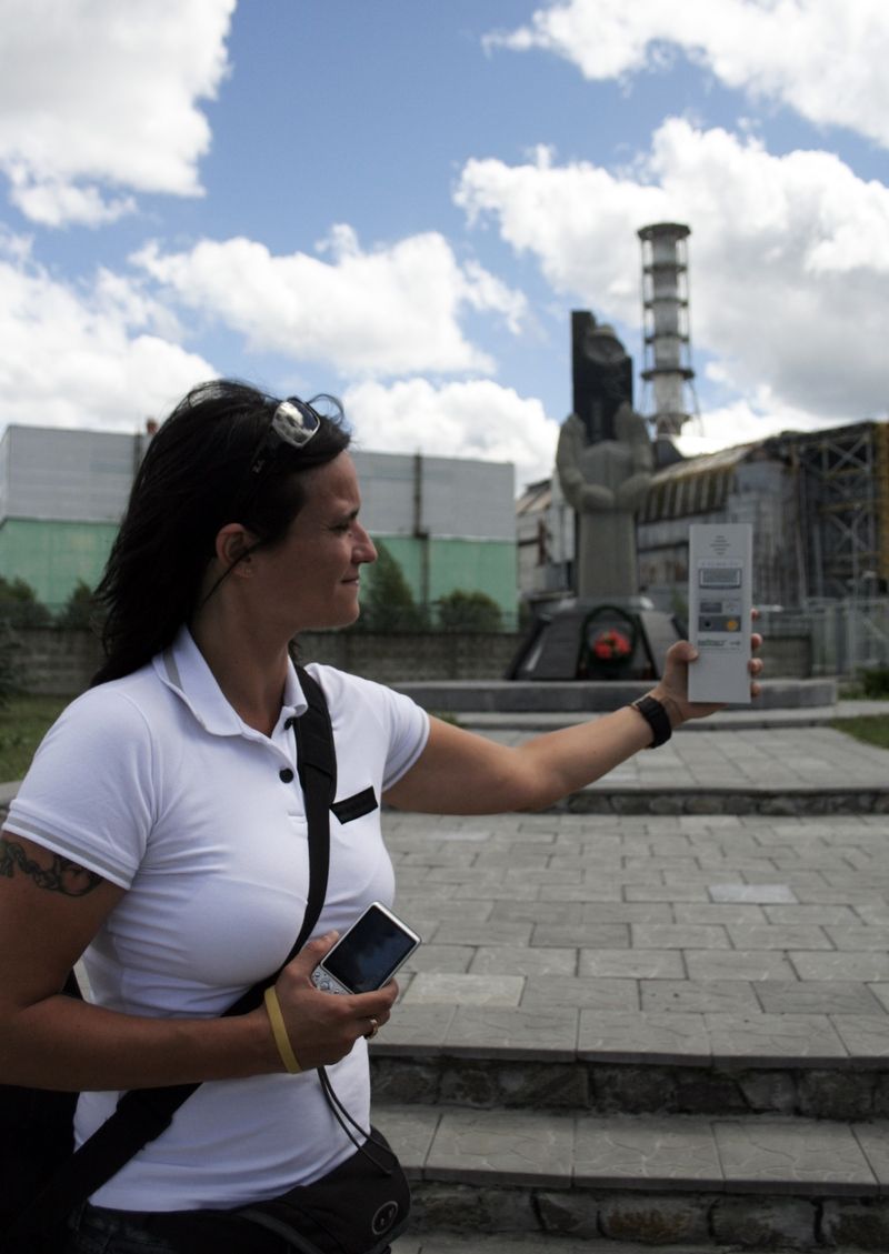 Účastnice expedice Simona stojí s dozimetrem před památníkem a 4. blokem jaderné elektrárny Černobyl.