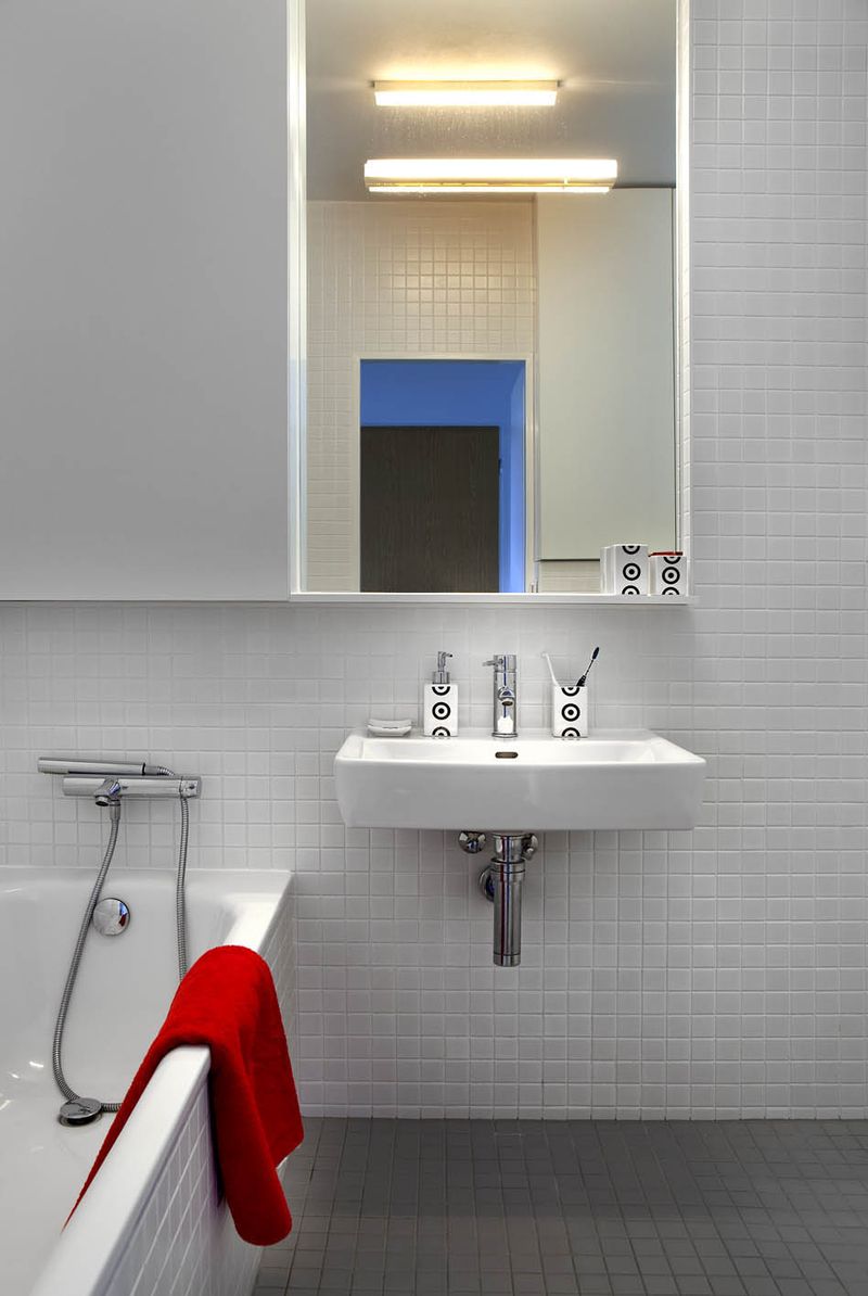 Koupelna je barevně laděná stejně jako celý byt, stěny jsou obloženy bílou keramikou a bílé sanitární předměty jsou doplněny úložnými skříňkami, prostor se zvětšuje velkým zrcadlem. 