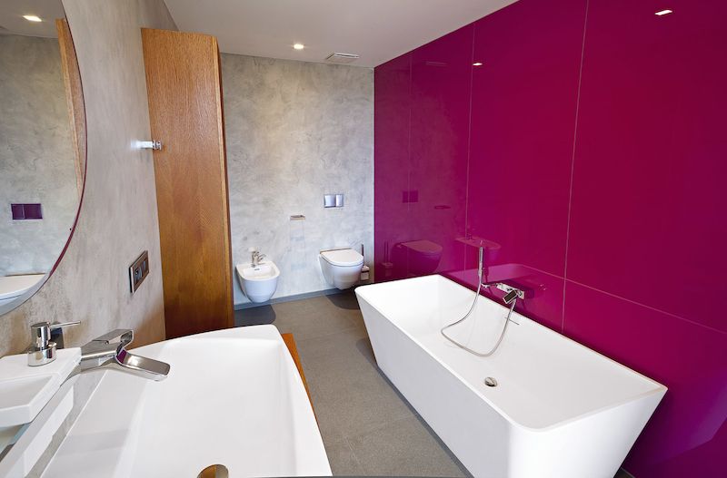 Každá z místností má svůj výrazný barevný akcent. V koupelně jsou to skleněné obklady v cyklámenově růžové, elegantně kontrastující s neutrálním základem cementové stěrky.