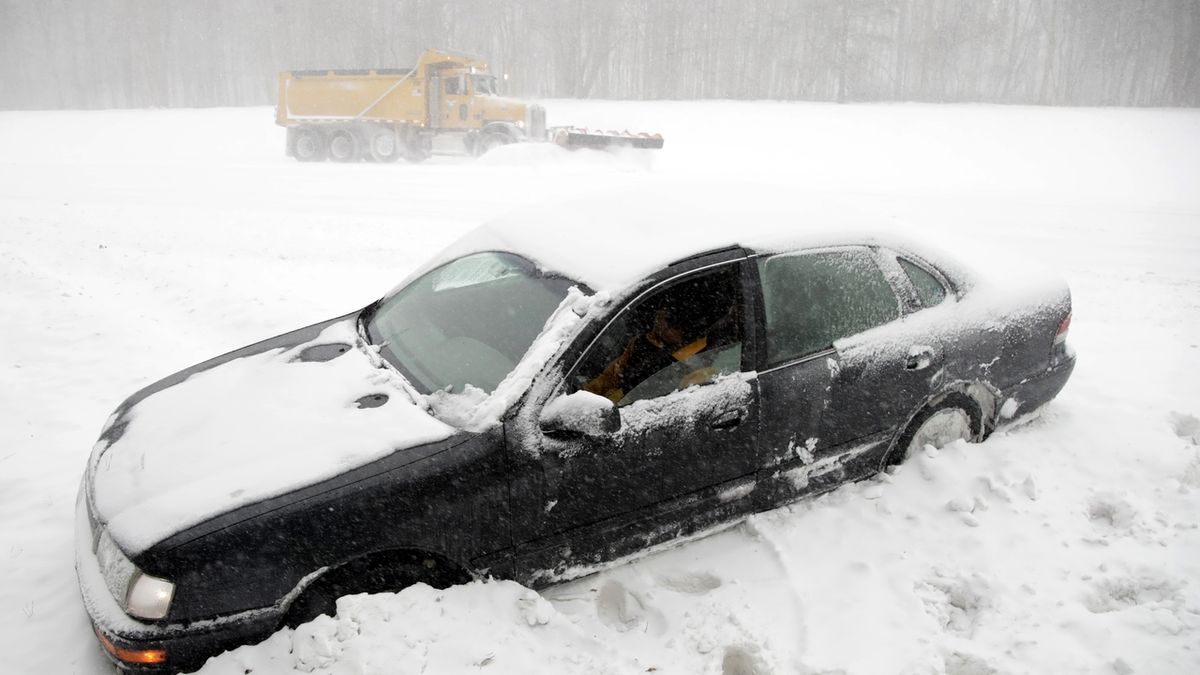 Řidič v New Jersey čeká, až ho vytáhne odtahovky, zatímco sněžný pluh prohrnuje silnici. 