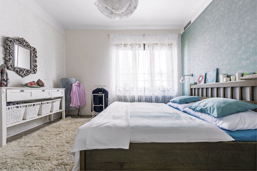 Ložnice je laděná do uklidňujících tónů modré a nadčasové bílé. Obrazy položené na čele postele a krajkové svítidlo jsou opět Moničinými výtvory.