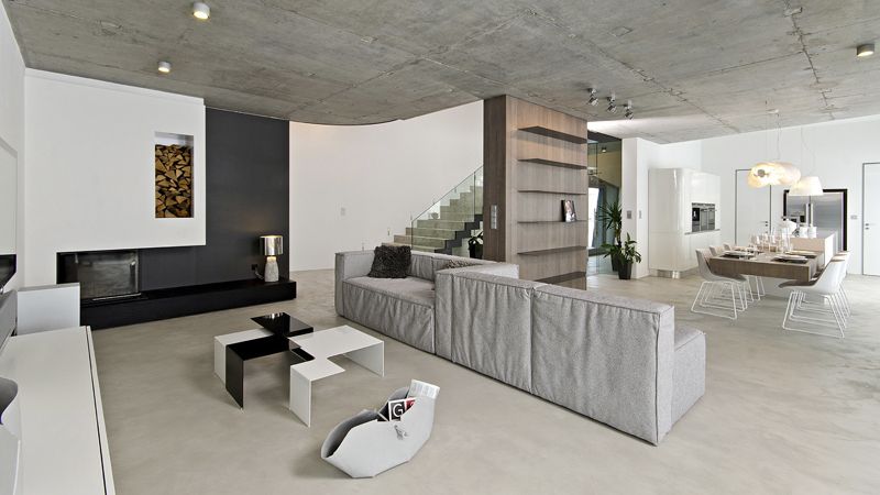 Společný obývací prostor se nese ve střízlivém duchu. Minimalistickou škálu barev provází minimum nábytku. Pohledový beton, cementovou stěrku a bíle lakované MDF desky doplňuje dubové dřevo (jídelní stůl, policová stěna) a čiré kalené sklo.  