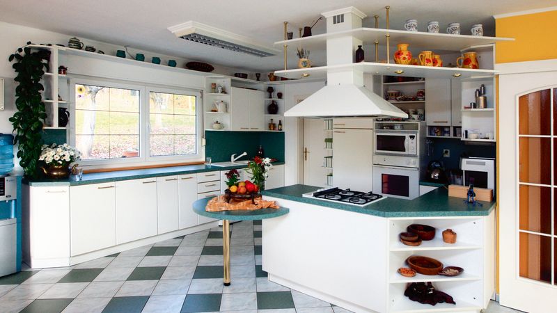 Bílo-zelenou kuchyň atypického tvaru doplňují červené a oranžové doplňky.