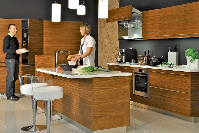 Kuchyně LINE v provedení dýha americký ořech, mat má jednoduché řešení moderní kuchyňské linky, praktické vysoké potravinové skříně, barové sezení (barová židle MIDJ – Bongo), osvětlení pracovní plochy je integrované do horních skříněk.