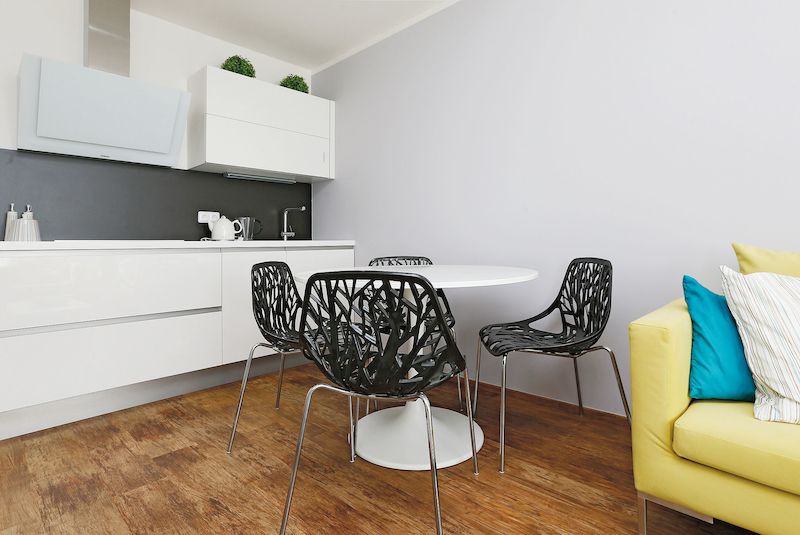 Bílý kulatý jídelní stůl doplňují černé plastové židle na kovových nožkách. Jídelna tvoří funkční předěl mezi kuchyní a relaxačním prostorem s televizí.
