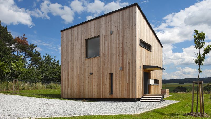 Kompaktní obdélníková stavba s pultovou střechou a fasádou obloženou neupraveným dřevem harmonicky ladí s prostředím. 