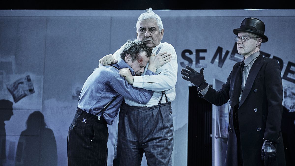 Willy Loman (Miroslav Donutil) v náručí se synem Biffem (Viktor Dvořák), přihlíží Willyho bratr Ben (Milan Kačmarčík).