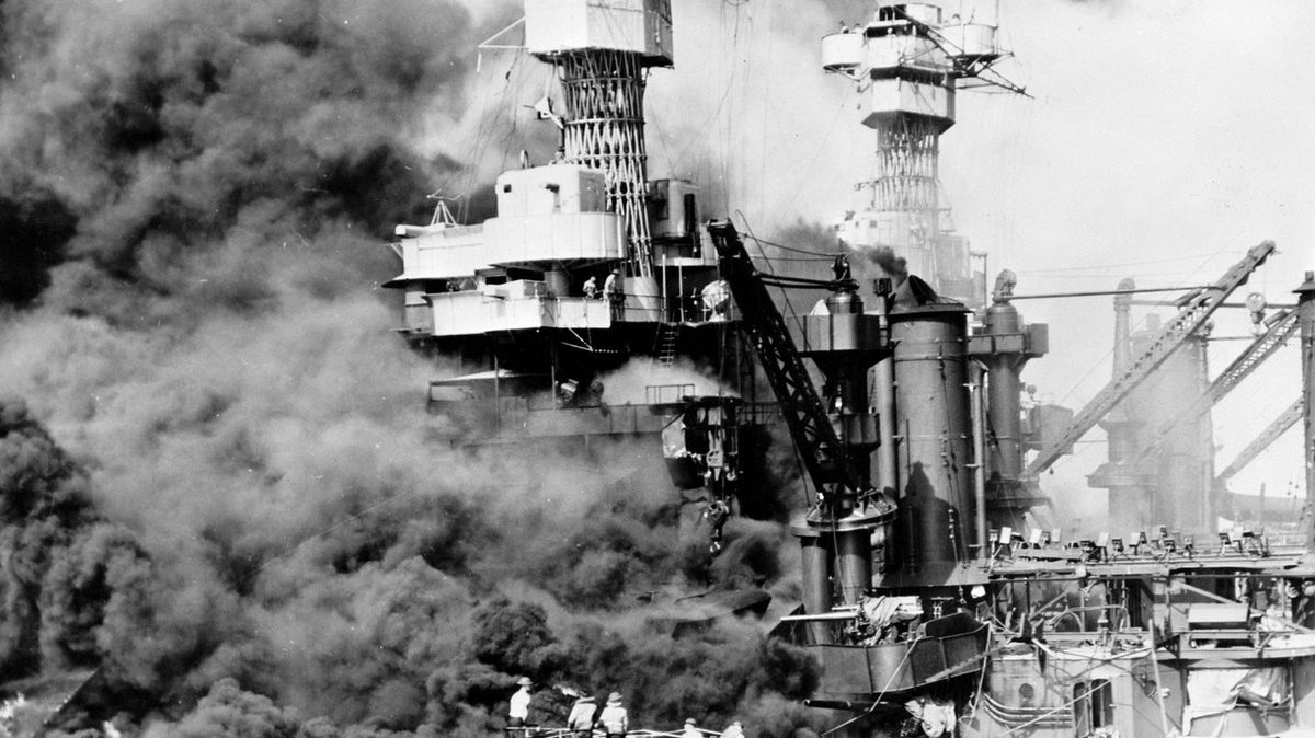 Bitevní loď USS Oklahoma se po zásahu převrátila, za ní hoří další lodě