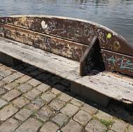 Dřevěná lavička na náplavce během sobotního dne. Všechny vypadají podobně, jedna  je na tom ale ještě hůř.