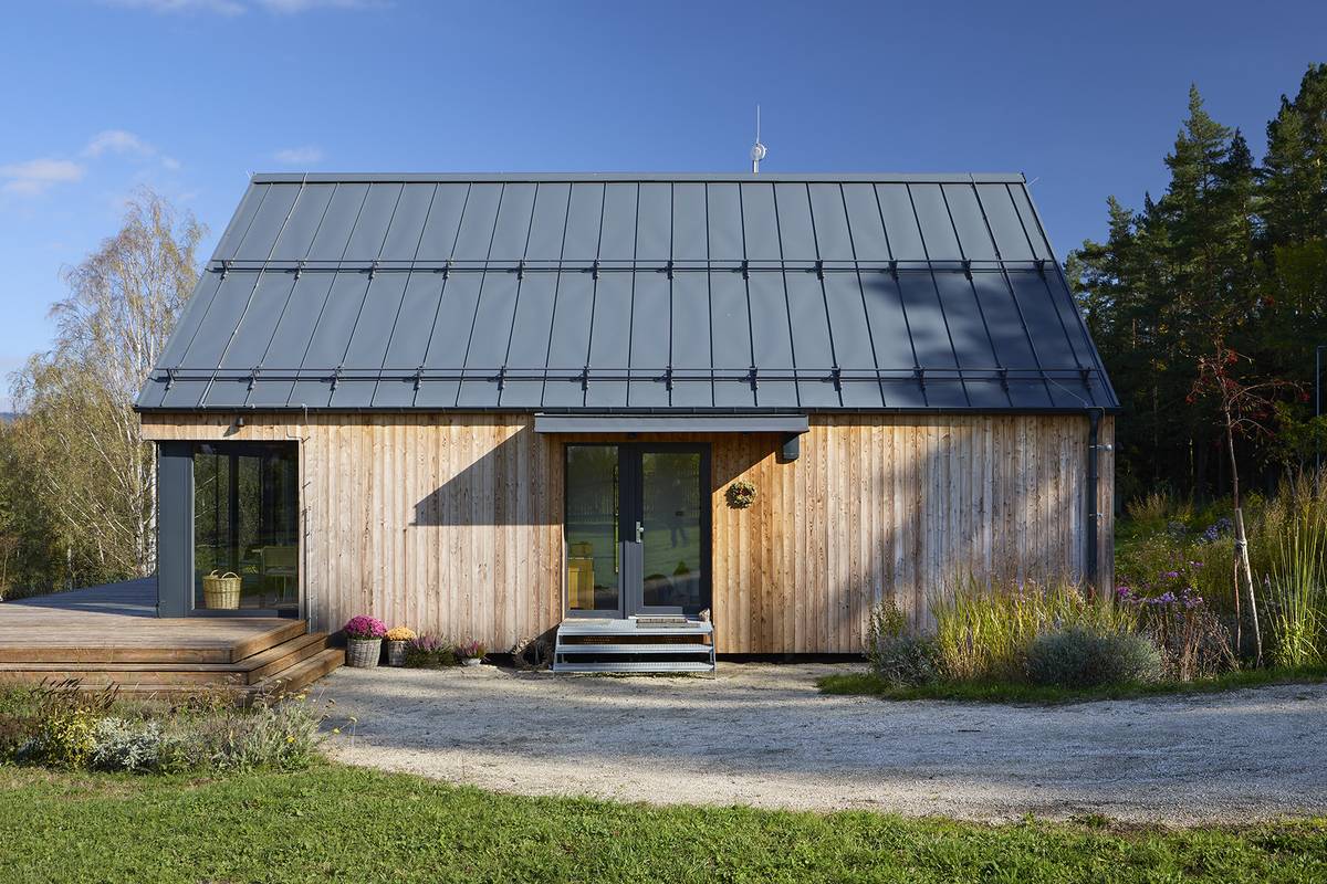 Pohled od příjezdové cesty – dům je založený na ocelových nosnících zakotvených do betonových patek. Od vjezdu na pozemek k němu vede světlým pískem vysypaná pěšina.