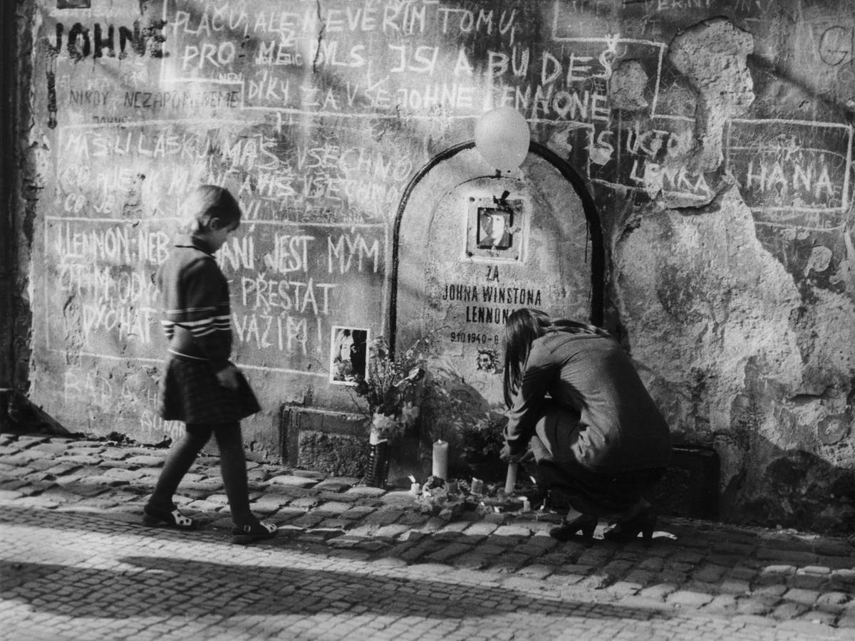 Symbolický náhrobek se na zdi na Velkopřevorském náměstí objevil už krátce po Lennonově smrti v roce 1980. Úřady nechaly zeď přetřít, ale upomínka se objevovala znovu a znovu. Tato je z roku 1985.
