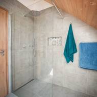 Moderní sprchový kout v podkroví kombinuje čistý design velkoformátových šedých obkladů s přírodním dřevem na stropě.